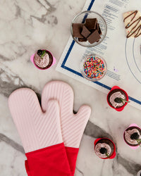 Top view of the Red Oven Mitts next to GIR Baking mat, next to some cupcakes in the Strawberry Swirl Cupcake Liners on a marble surface. 