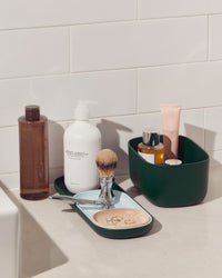 A Small Dark Green Storages Bins with beauty products in it next to a Dark Green Nesting Tray on a white background.