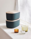Two Small Dark Green Storages Bins with wooden lids stacked on top of each other on a cream background.