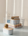Two Small Light Blue Storages Bins with wooden lids with a Large Light Blue Wire Basket on a cream background.