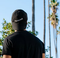 The back of a man wearing the The Thinking Cap in black with palm trees in front of him