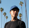 A man wearing Thinking Cap in Cobalt with palm trees behind him. 