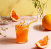 A straw from the Glass Straws in Warm Set in a glass of orange juice with flowers garnishing it and oranges in the background. 