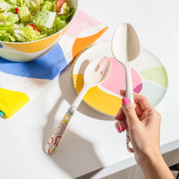 A hand holding the Bamboo spoon with the Bamboo Fork resting on a Poketo Bamboo Salad plate Plate with a salad in the top left corner.